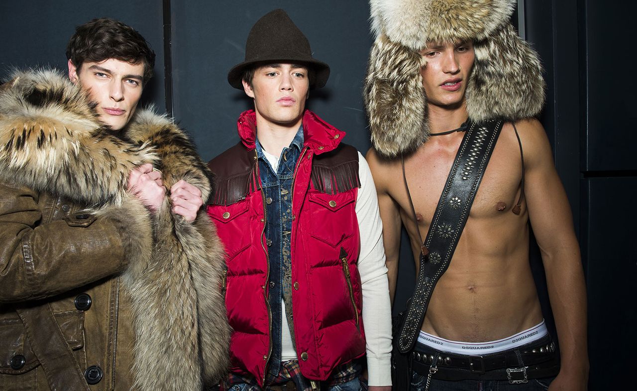 Three male models wearing looks from Dsquared2&#039;s collection. One model is wearing a brown coat with fur. Another model is wearing a dark hat, white long sleeve top, sleeveless denim jacket and red gilet with brown fringe on top. And the last model is wearing an oversized fur hat, jeans and is bare chested