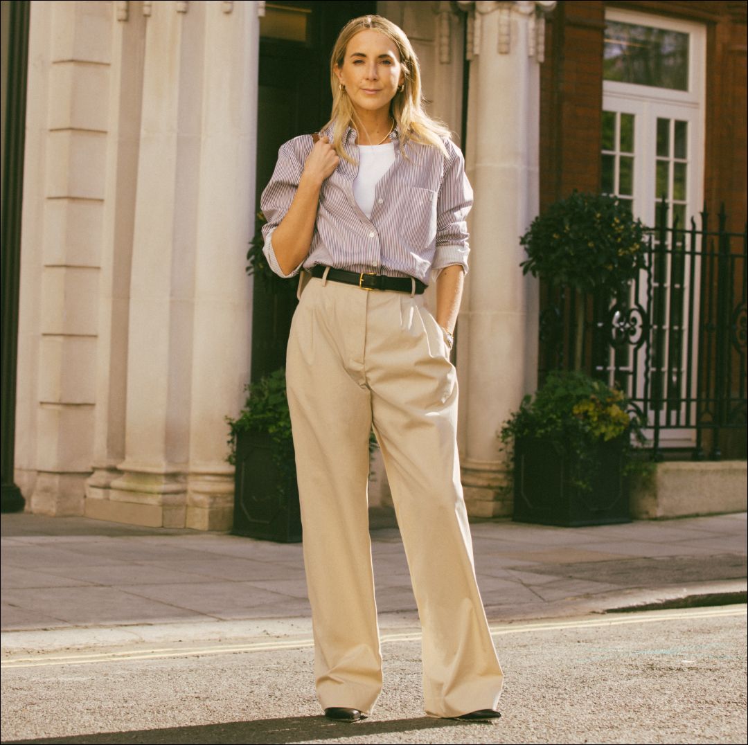 Model wears high-waisted tan pants, black belt, striped button-down shirt, and white tank top and is standing in front of a white building.