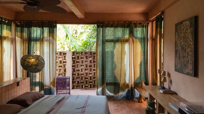 Tropical bedroom interiors, with colorful curtains, terracotta floors and walls, pendant lights, sculptural wooden furniture, and lush views.