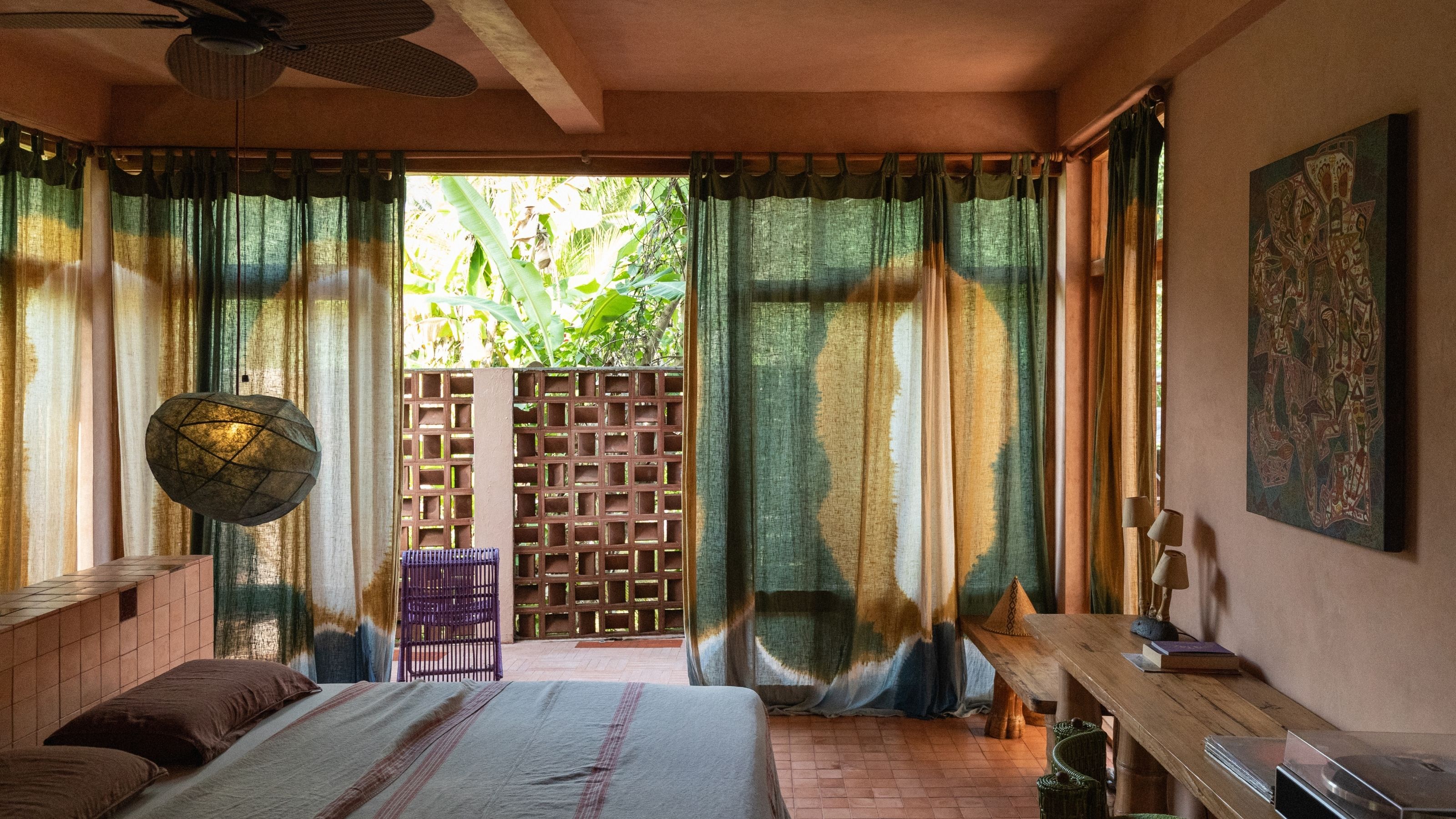 Tropical bedroom interiors, with colorful curtains, terracotta floors and walls, pendant lights, sculptural wooden furniture, and lush views.