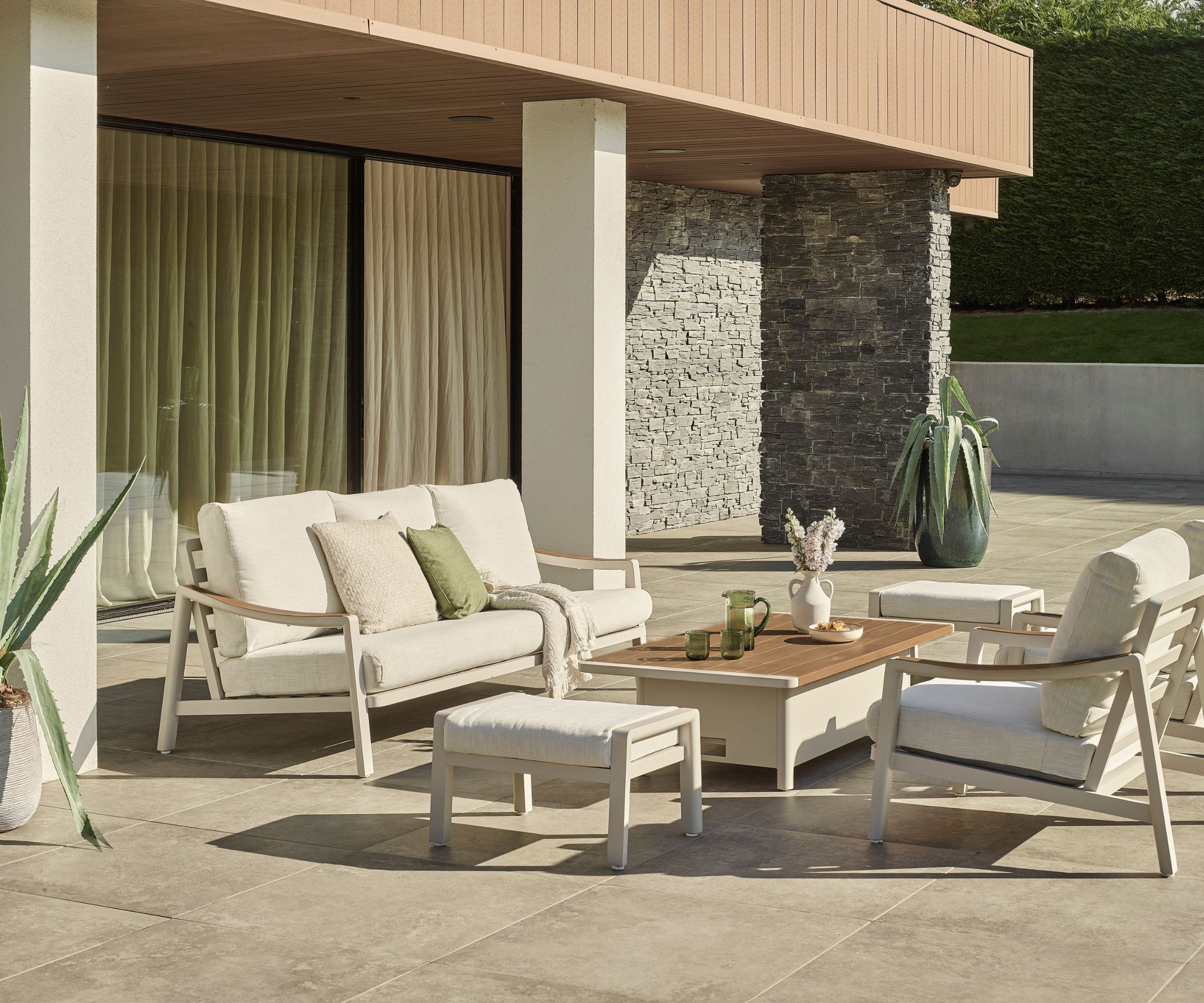Cream outdoor sofa and armchairs around wooden coffee table on stone patio, stacked stone pillar behind.