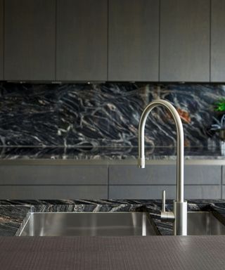 Close up of black and white marbled worktop with metal sink and mixer tap and dark grey kitchen cabinets.