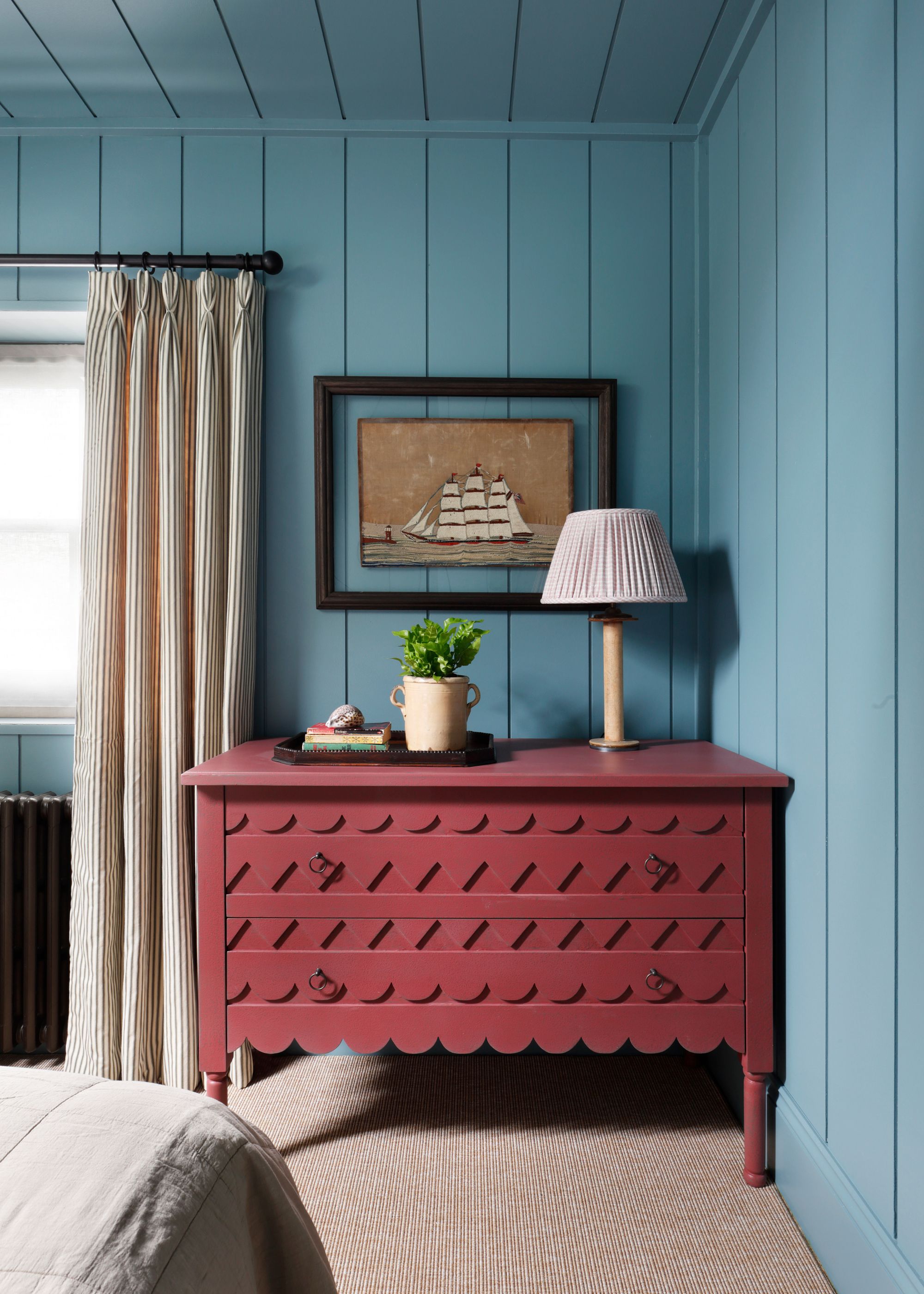 A bedroom with blue wall paneling on the walls and on the ceiling. A raspberry red chest of drawers with a lamp and a plant pot.
