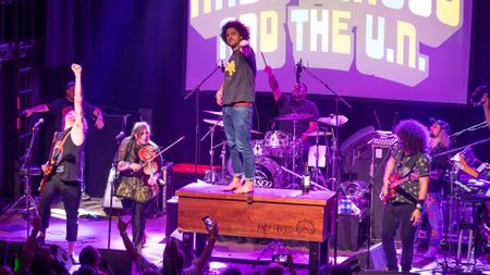 A rock band on stage. One person is standing on top of a brown box and there are guitarists and a violinist nearby.