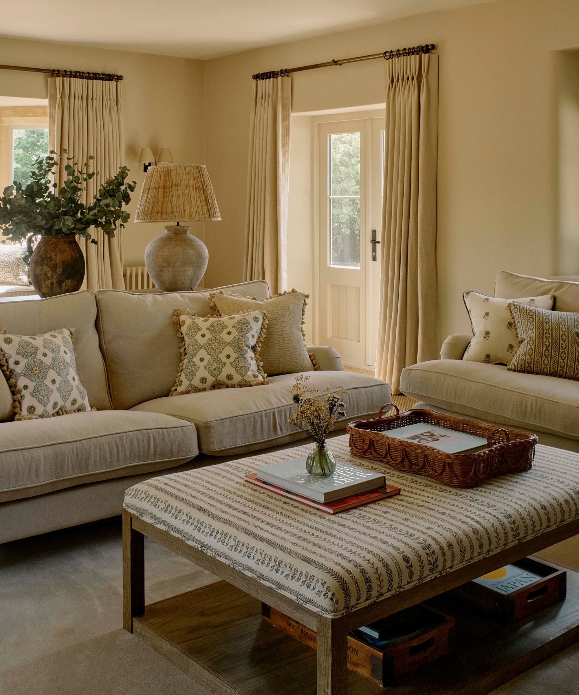 warm cozy soft neutral living room with french doors out to the garden, two beige couches styled with patterned pillows with statement trim edging and a floral and wooden upholstered ottoman with piping trim