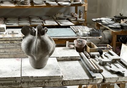 A gray ceramic vase by Pierre Casenove shown in his ceramics studio in Jura on a table with ceramics samples and shelves with prototypes in the background