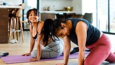 Two woman on yoga mats in a home look at each other and smile