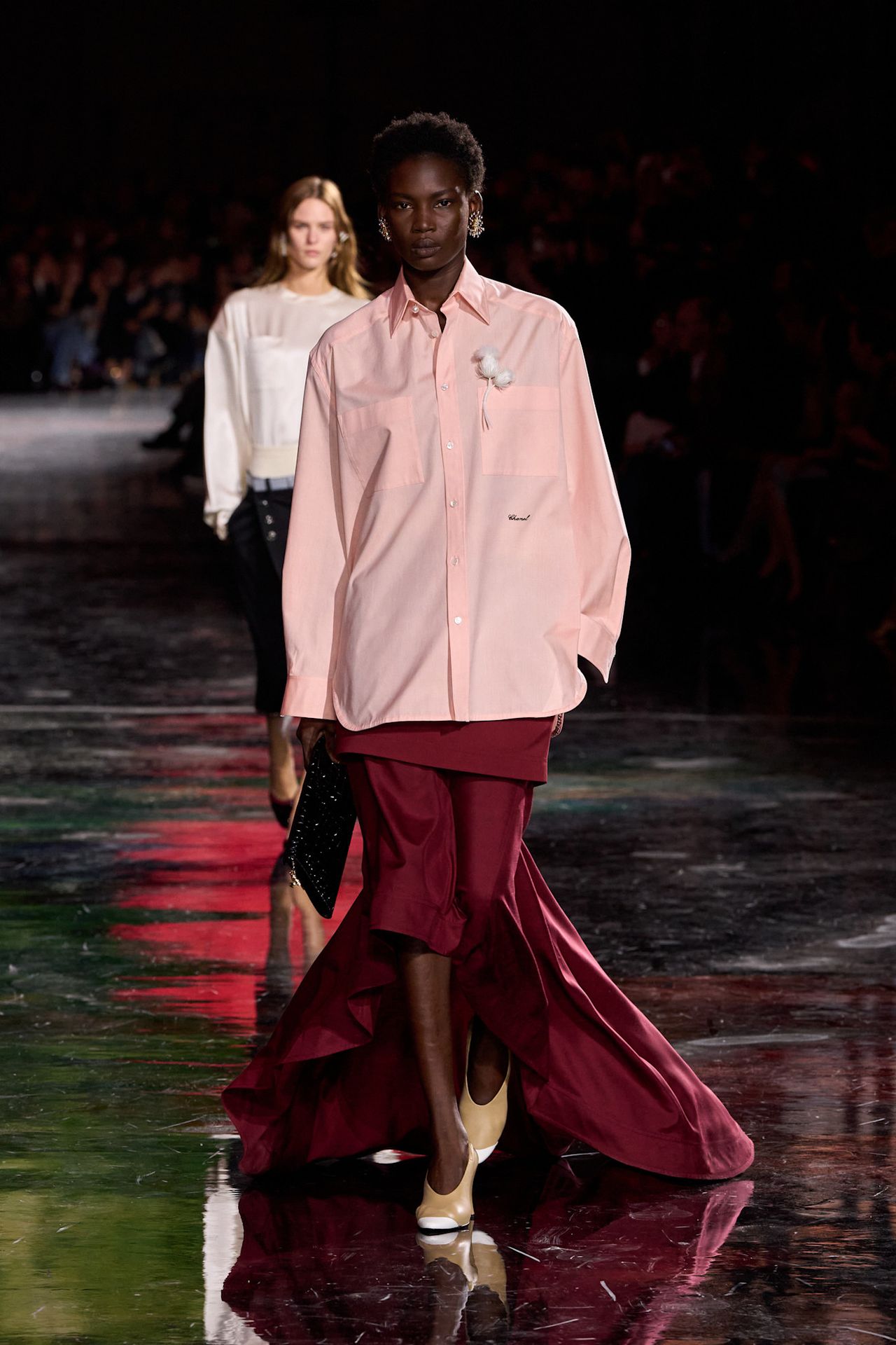 A Chanel model wearing an oversize pink button-down shirt, a burgundy high-low skirt styled with a floral brooch, a black clutch bag, and a pair of two-tone heels.