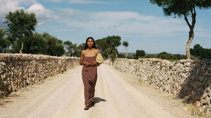 Woman wearing a Faithfull x Monikh brown maxi dress in the middle of the road, walking towards the camera