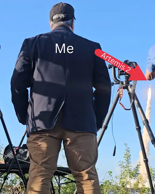 A man stands next to a camera as a rocket launches in the distance.
