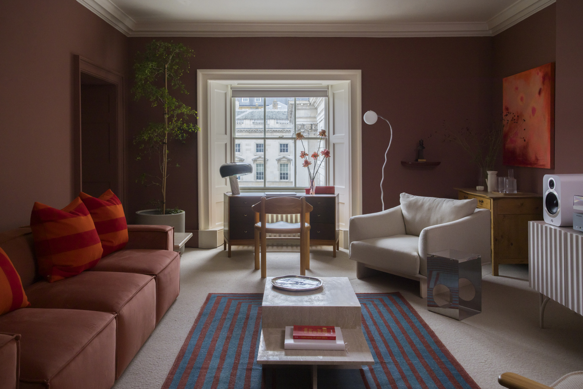 red living room with red sofa, red and orange cushion, blue and red striped rug, desk under a window, white armchair in front of a bar with artwork above and console table with speaker