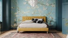 Pale blue wallpaper with white blossoms in a blue color drenched bedroom with wooden floors, pale red rug, low profile yellow modern bed with button back headboard