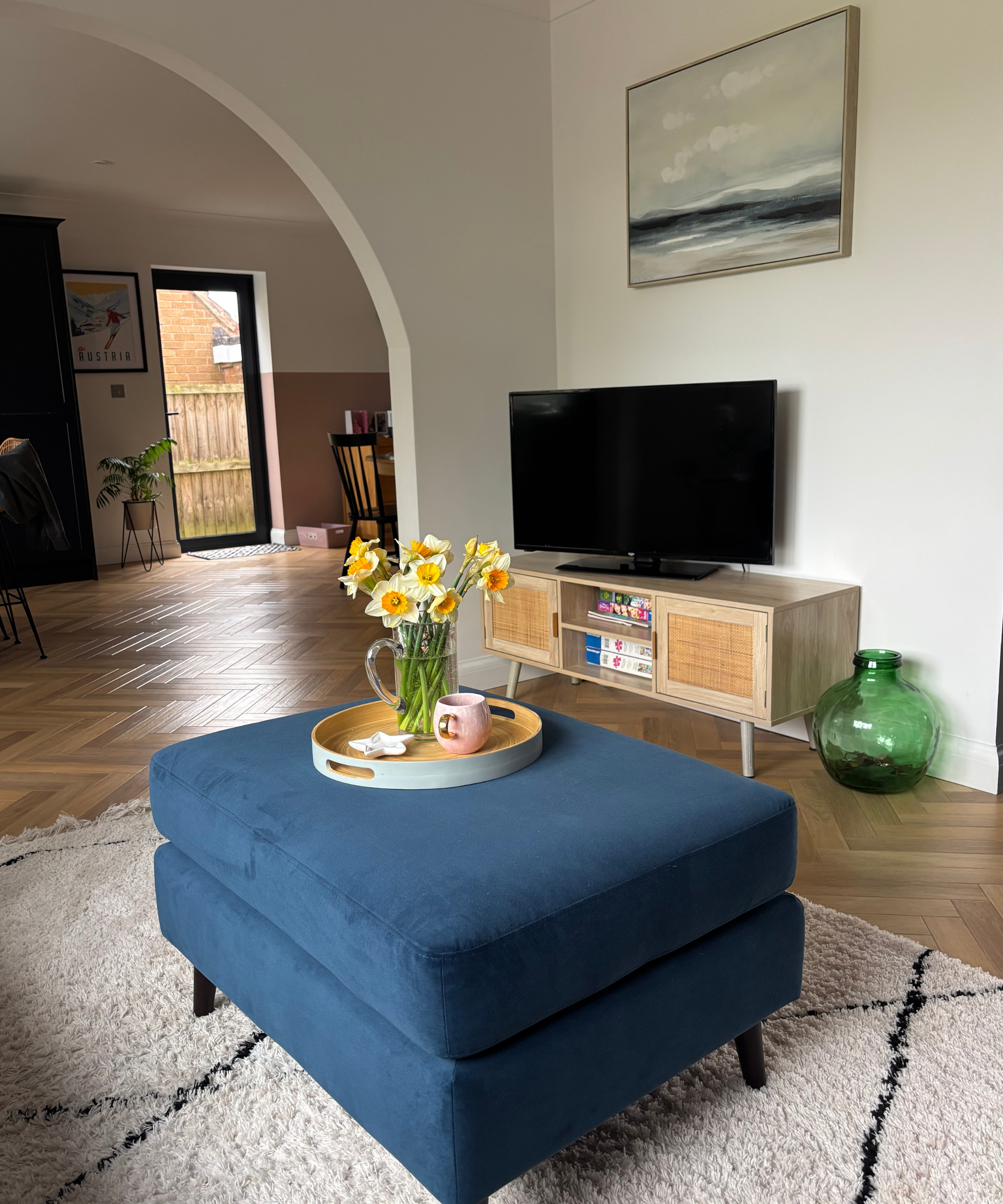 Laura's living room featuring a rattan-style TV unit, a painting, a blue poof with a tray of flowers and a mug on it, and a black and white rug underneath