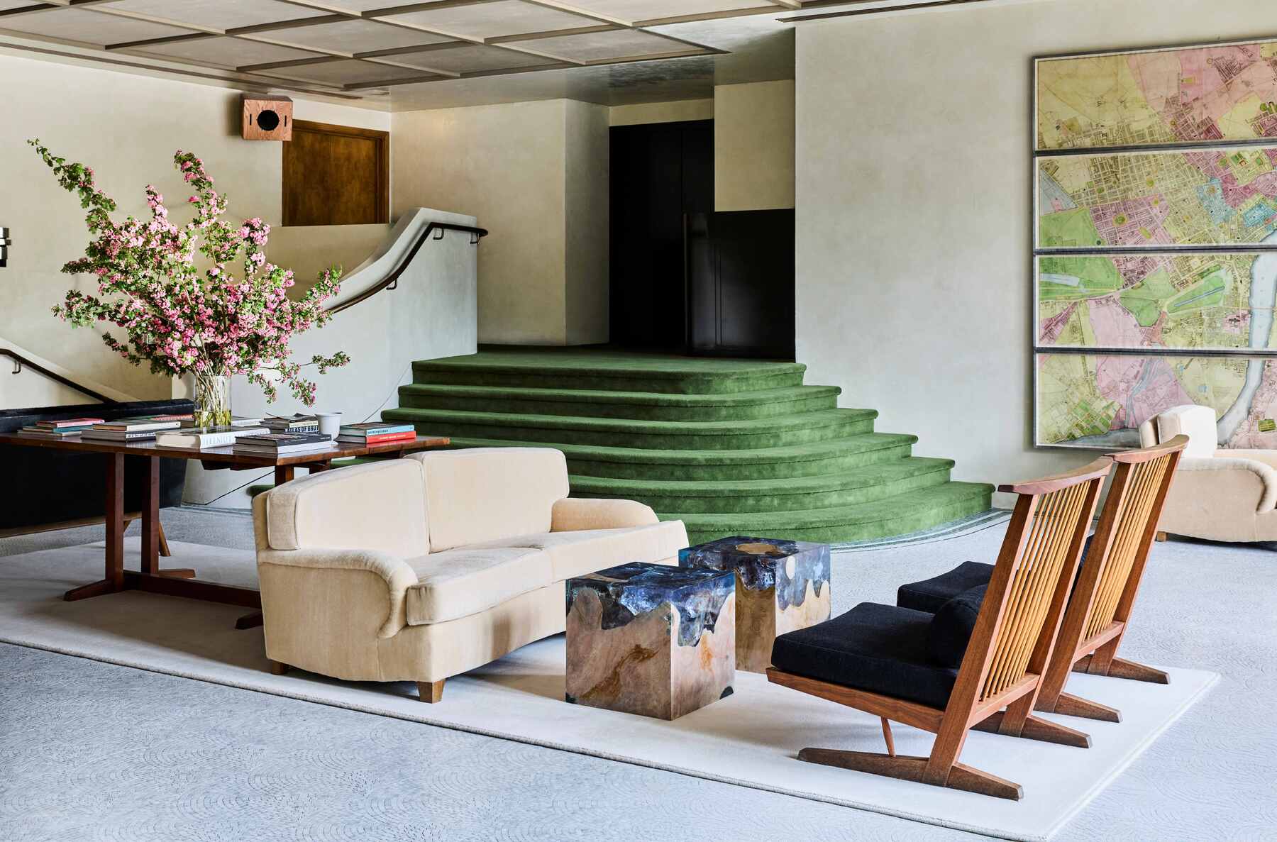 The lobby of a hotel, with velvet green staircases, vintage maps framed on the wall, and mid-century modern furniture in black velvet, wood, and beige fabric.