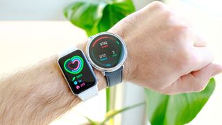 The Samsung Galaxy Fit3 on a user's wrist next to the Amazfit Active 2 with each device showing a step-count total for the day; a green plant is out of focus behind the watches