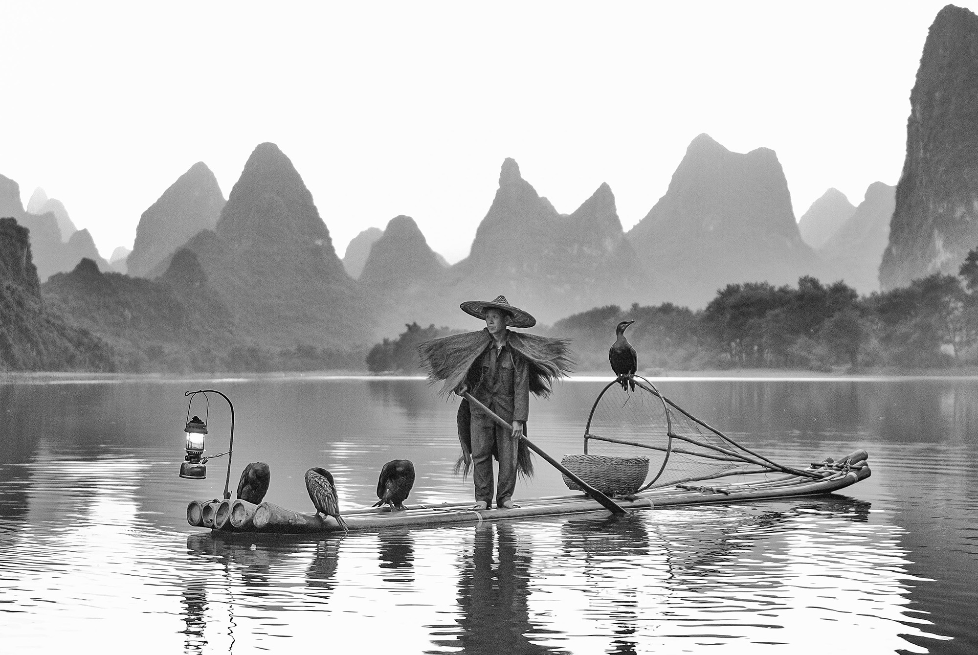 Black and white travel portrait of cormorant fisherman on lake against backdrop of hills