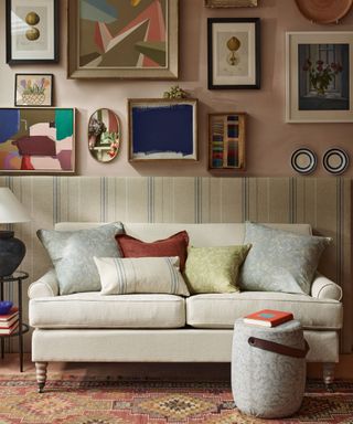 Living room with striped fabric paneling, pale pink painted walls with gallery wall or framed pictures and decorative objects, colorful rug and rounded footstool, cream two seater sofa with colorful scatter cushions