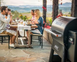 wood pellet grill on a patio with people eating at a dining table in the background