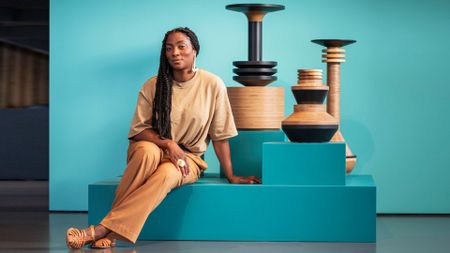 Simone Brester wears a terracotta-hued outfit while sitting next to some of her wooden creations in a seafoam green gallery space.