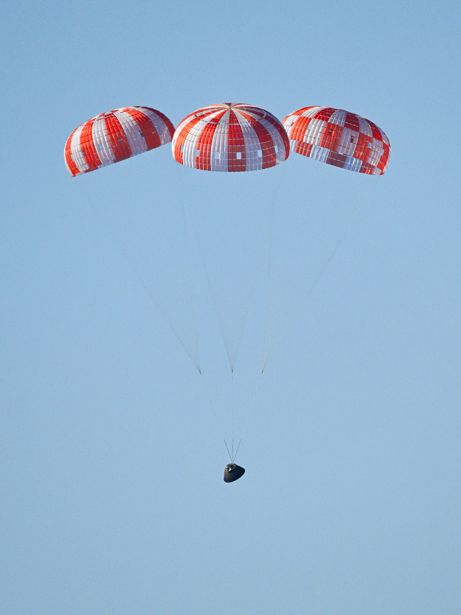 The Artemis 2 capsule is pictured falling throught a pale blue sky under three deployed parachutes.