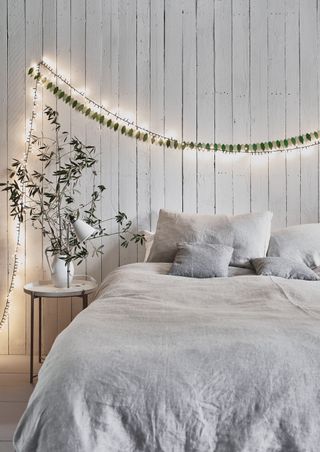 A bedroom with white shiplap decor, gray bedding, small round side table, green leaf bunting and string of fairy lights