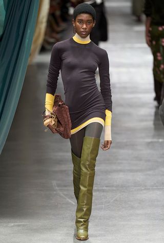 a photo showing some of the best thigh-high boots with a model walking the Fendi runway show wearing a yellow turtleneck under a gray knit dress styled with gray tights, green thigh-high boots, and a brown bag