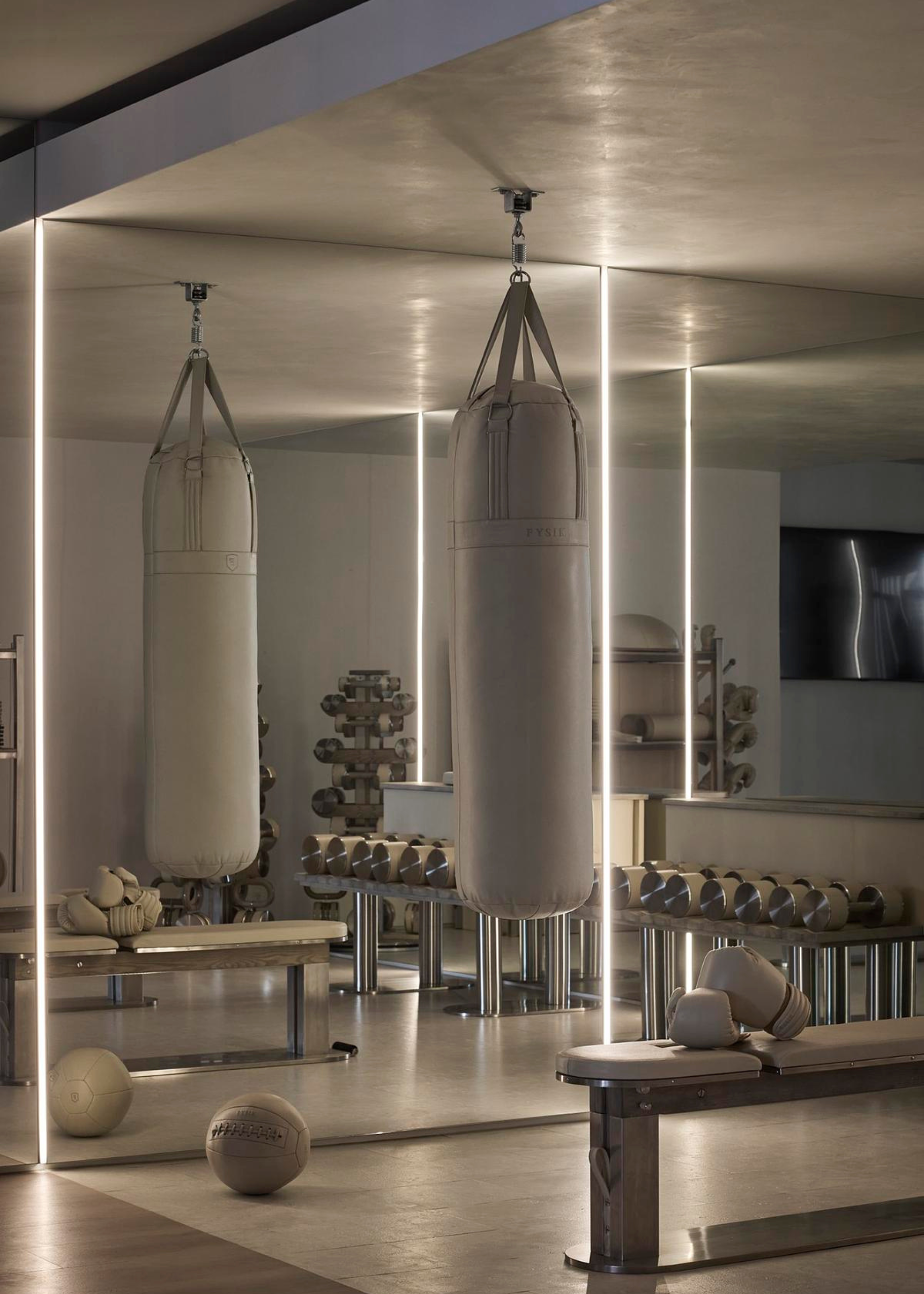 A home gym with beige walls, chrome accessories, a beige bench, a hanging punching bag, weighted balls, and a set of dumbells