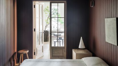A sleek and minimalist modern bedroom with paneled walls, beige bedding tucked under the pillows and double doors with paneled glass opening up to a dining room beyond with a large potted indoor tree