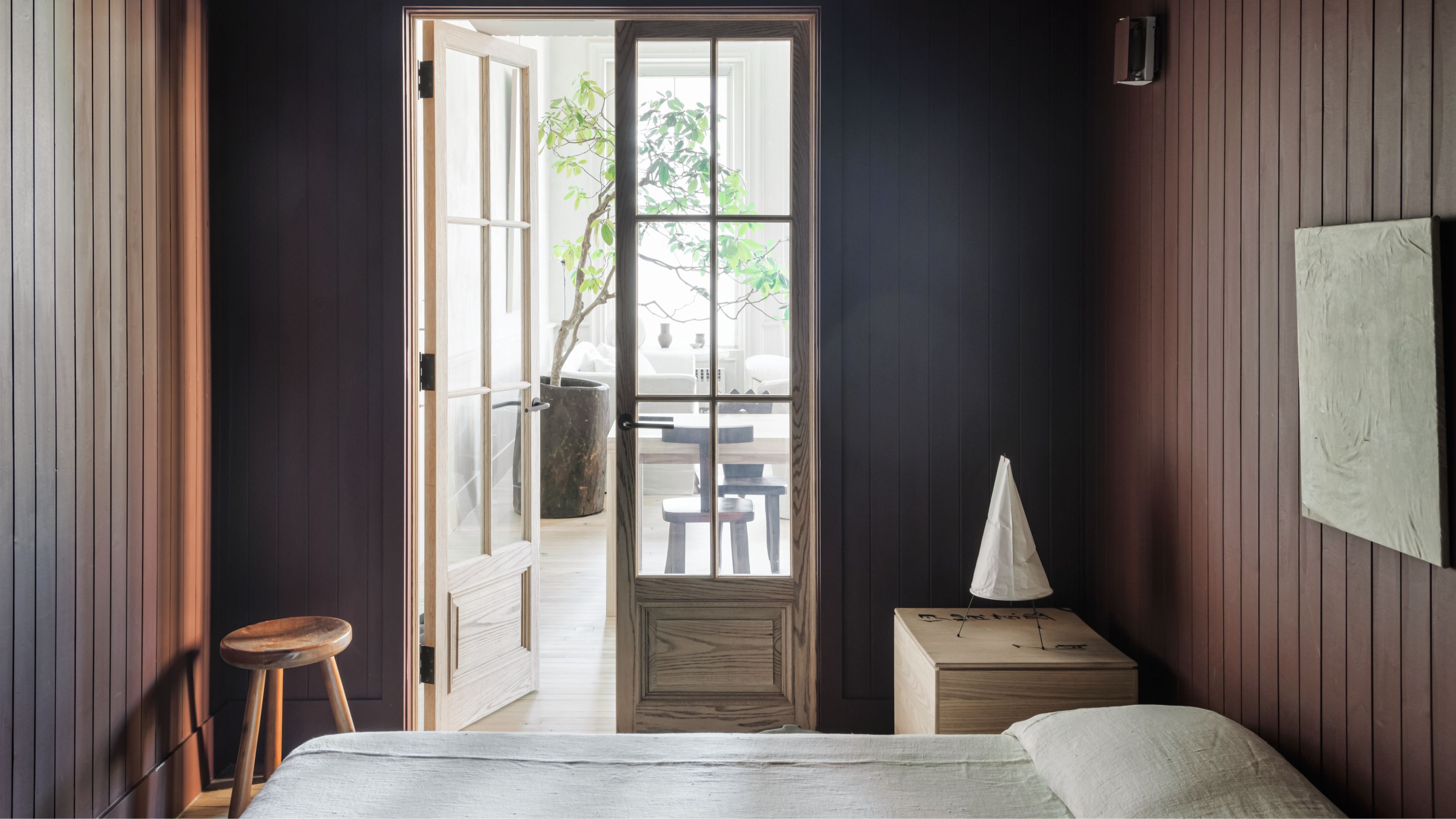 A sleek and minimalist modern bedroom with paneled walls, beige bedding tucked under the pillows and double doors with paneled glass opening up to a dining room beyond with a large potted indoor tree