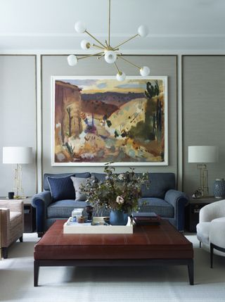 A large oil painting on a pale gray wood panelled wall above a blue sofa and leather ottoman, illustrating small apartment living room ideas.