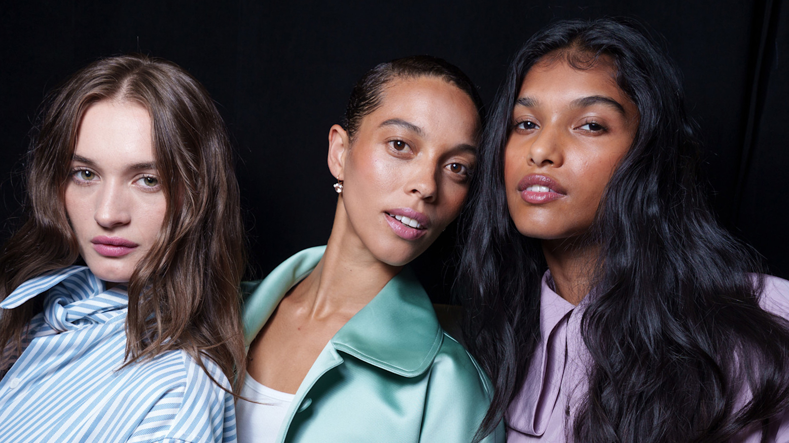 A trio of models with glossy hair stand backstage at fashion week.