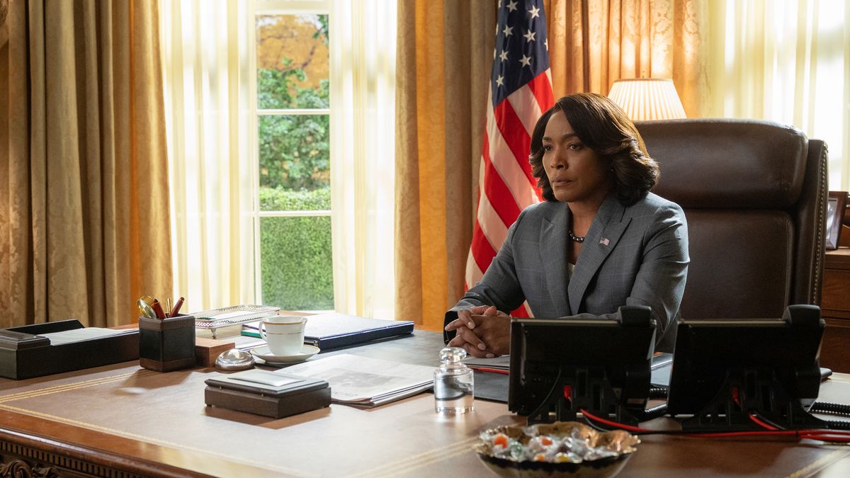 Angela Bassett as President Mitchell, sitting at her desk in the Oval Office wearing a grey suit and looking serious and thoughtful. 