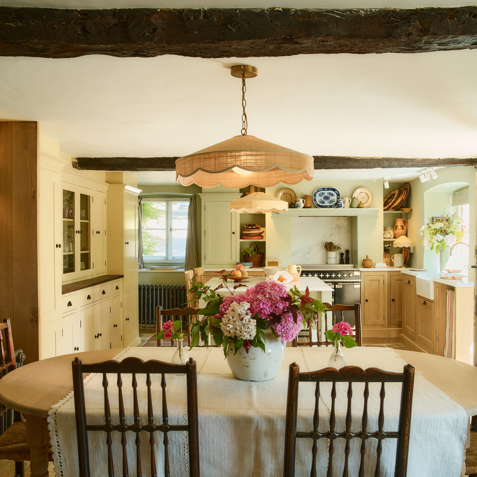 close up of oval kitchen table with wooden chairs and pink and white hydrangeas in a jug in the centre behind is a country style kitchen with cabinetry the ceiling has a large original beams