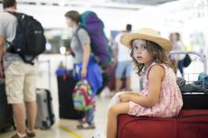 little girl with straw hat sitting on a suitcase at the airport. during check in at the airport the girl takes a seat on the luggage carrier. She looks tired from waiting.