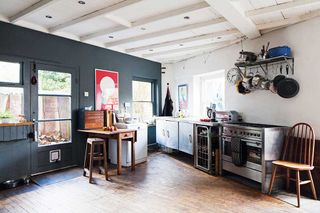 a freestanding kitchen in a converted brewery - with stainless steel units and oven to the right, a hanging pot rack on the wall, a black wall to the left with a butcher's block and a small fridge, and white wooden beams in the ceiling