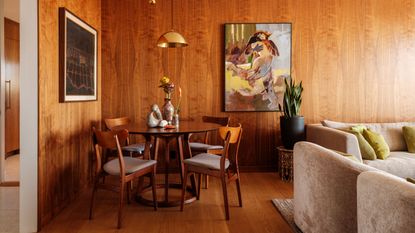 A living room with wooden flooring and wooden walls with a sitting area off to the right side that has two beige sofas. There is a dining area in the main view that has a round wooden table and four matching chairs.