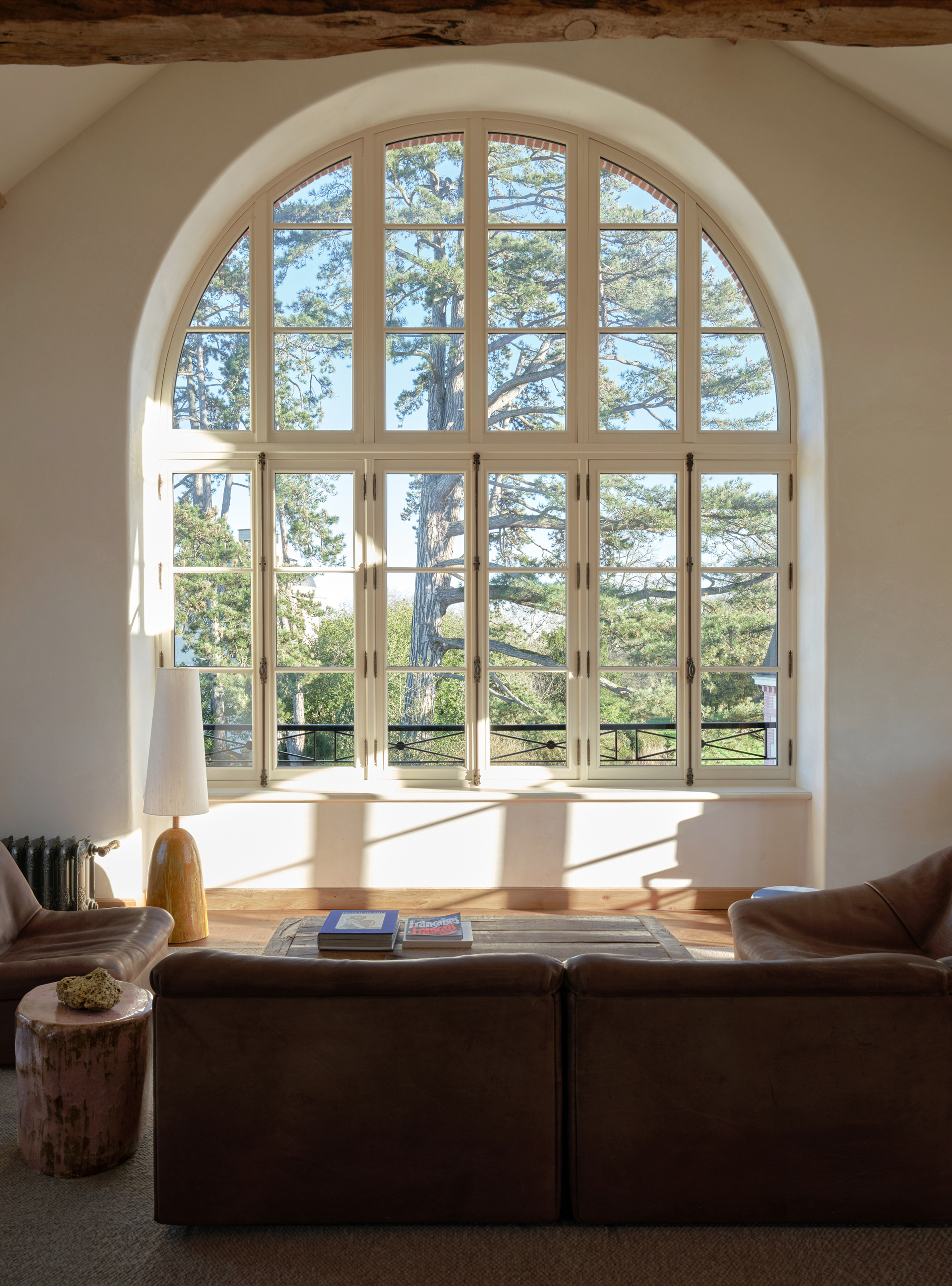 A modern rustic hotel room filled with sunshine, leather, and wooden furniture features an arched window looking out to greenery.