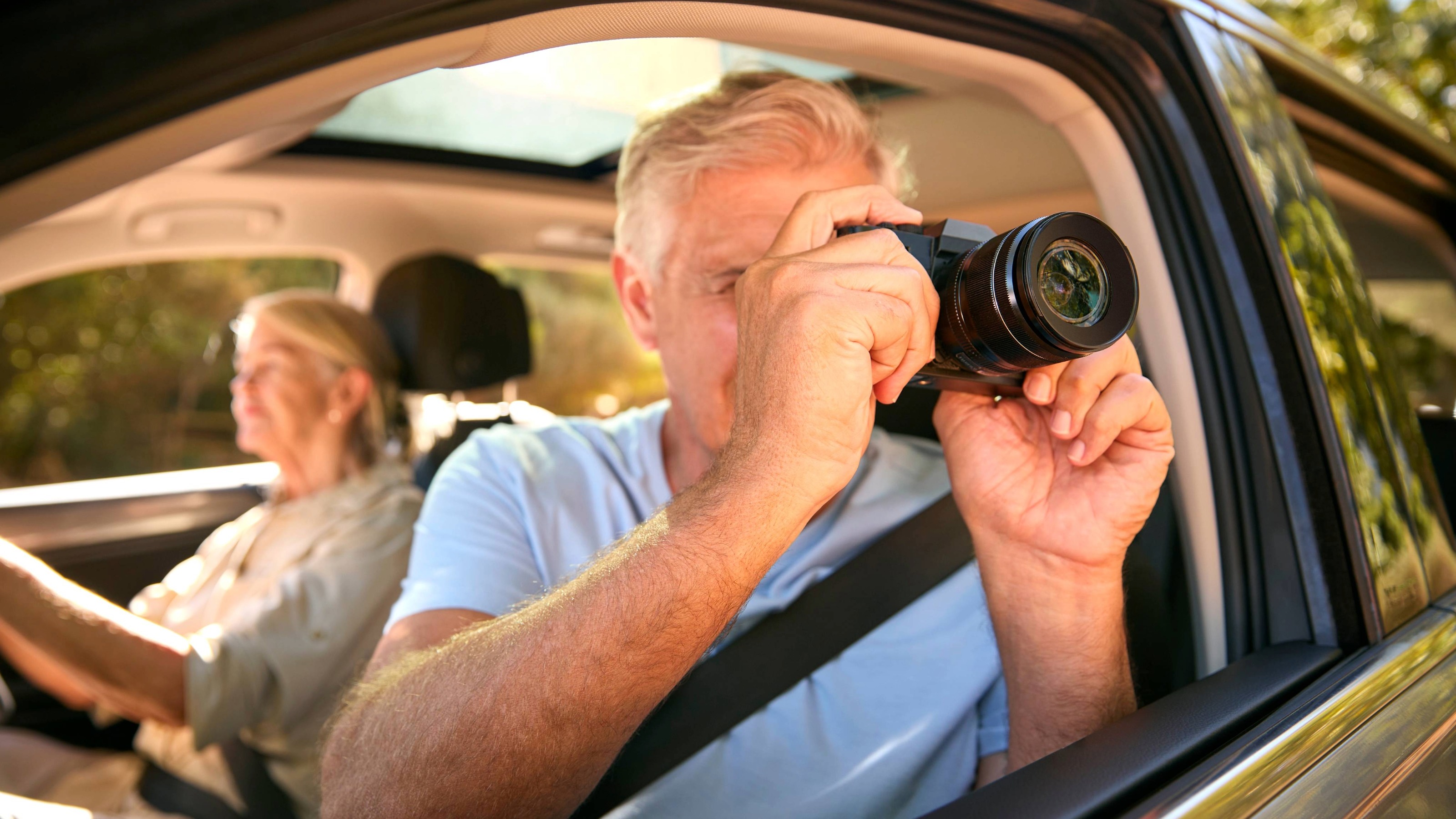 Male Passenger Taking Photo With Camera As Senior Couple Enjoy Day Trip Out In Car