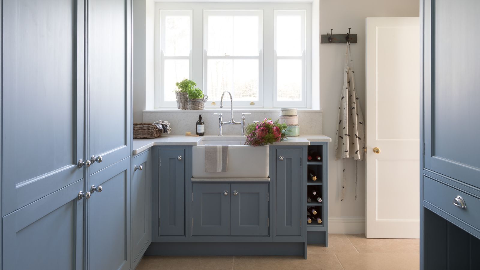 A utility room with blue built-in cabinets and a white farmhouse sink beneath a large framed window. 