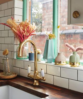 A brass faucet/tap in kitchen with white tile decor and assortment of pastel-colored planters and vases sitting on ledge, with teal green window frame