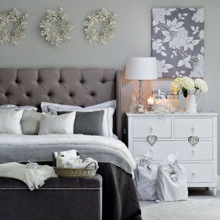 Pale grey bedroom, double bed, grey bedlinen, white bedside table chest of drawers, floral canvas, gift bags. white flower wreath feature display
