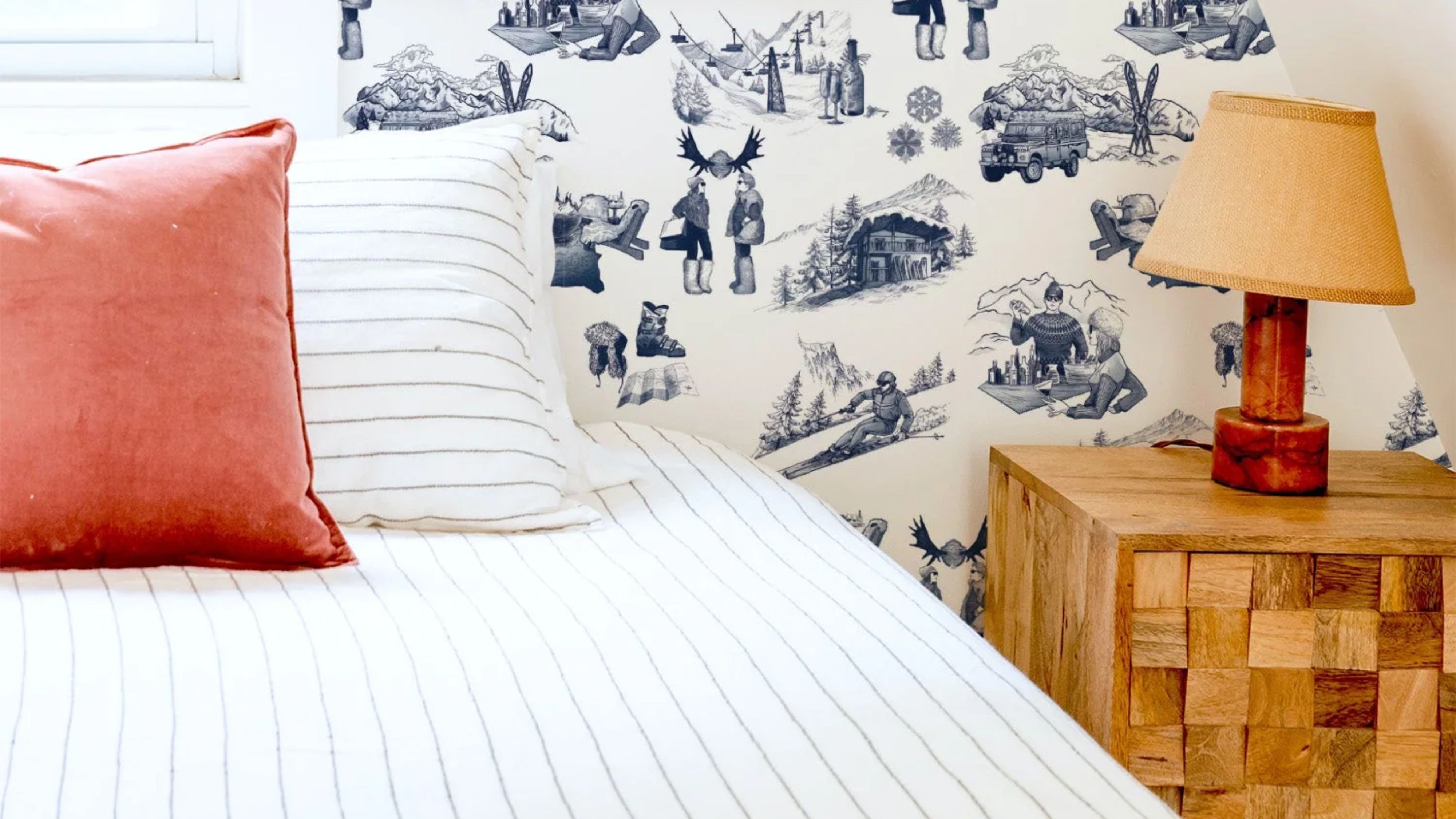 Bright bedroom featuring winter toile patterned wallpaper, a natural wood bedside table, and a bed styled with pinstripe bed linens