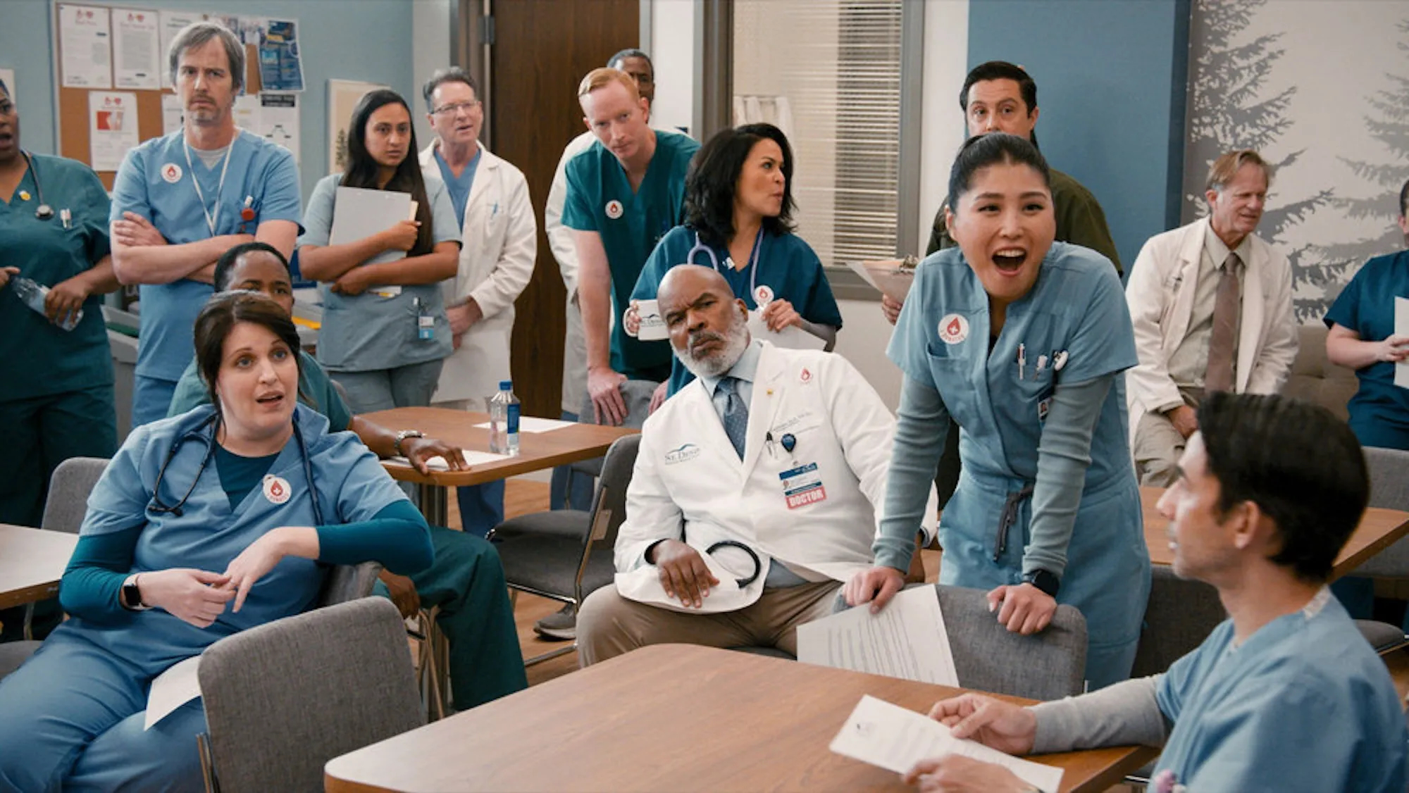 A group of doctors and nurses sit and stand in a crowded staff lounge in a scene from 'St. Denis Medical'