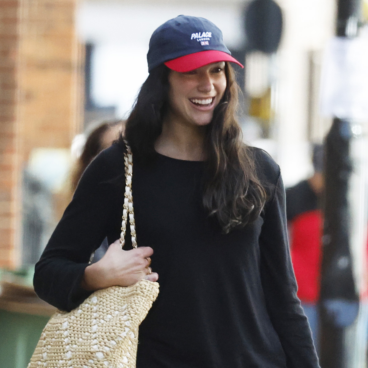 Dua Lipa wearing a two-tone baseball cap with a black long-sleeve T-shirt, white jeans, white sneakers, and a raffia tote at the airport in London