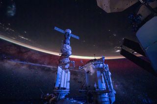 two pillars formed by space station modules sprout from the bottom, draped in mechanical accessories, with a swath of milky as a backdrop and the curvature of the earth hanging from above.