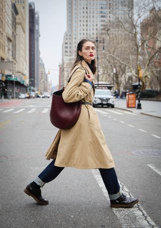 A woman crosses the street in New York City wearing jeans, a trench coat and black Oxford shoes.