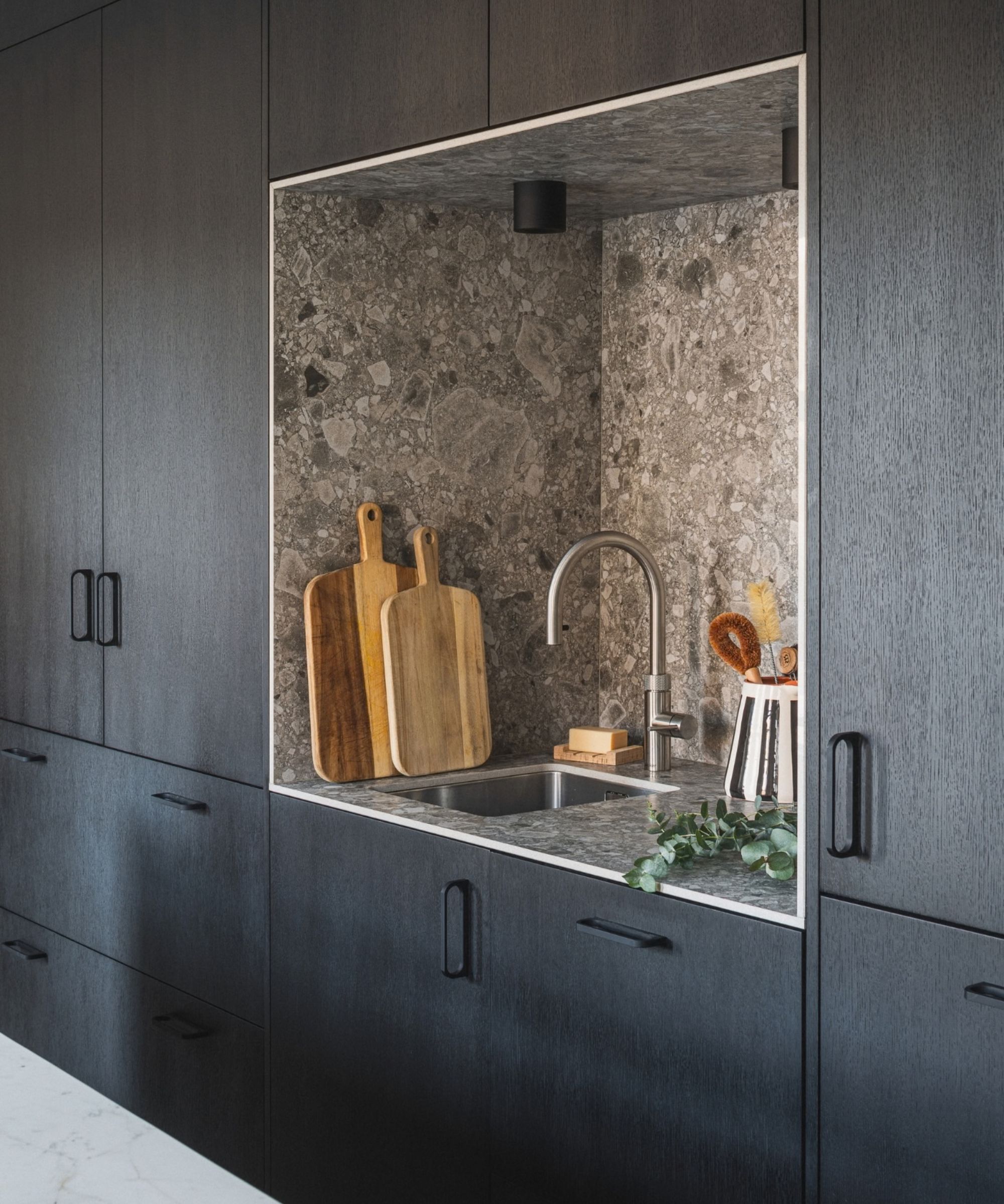 A dramatic kitchen featuring floor-to-ceiling dark charcoal grey oak cabinetry with matte black handle hardware. A recessed niche is cut into the cabinetry, with walls and worktop clad in grey textured stone with a fossil-like pattern. Inside the niche, a stainless steel undermount sink sits beneath a brushed nickel gooseneck tap, with two wooden chopping boards leaning against the back wall