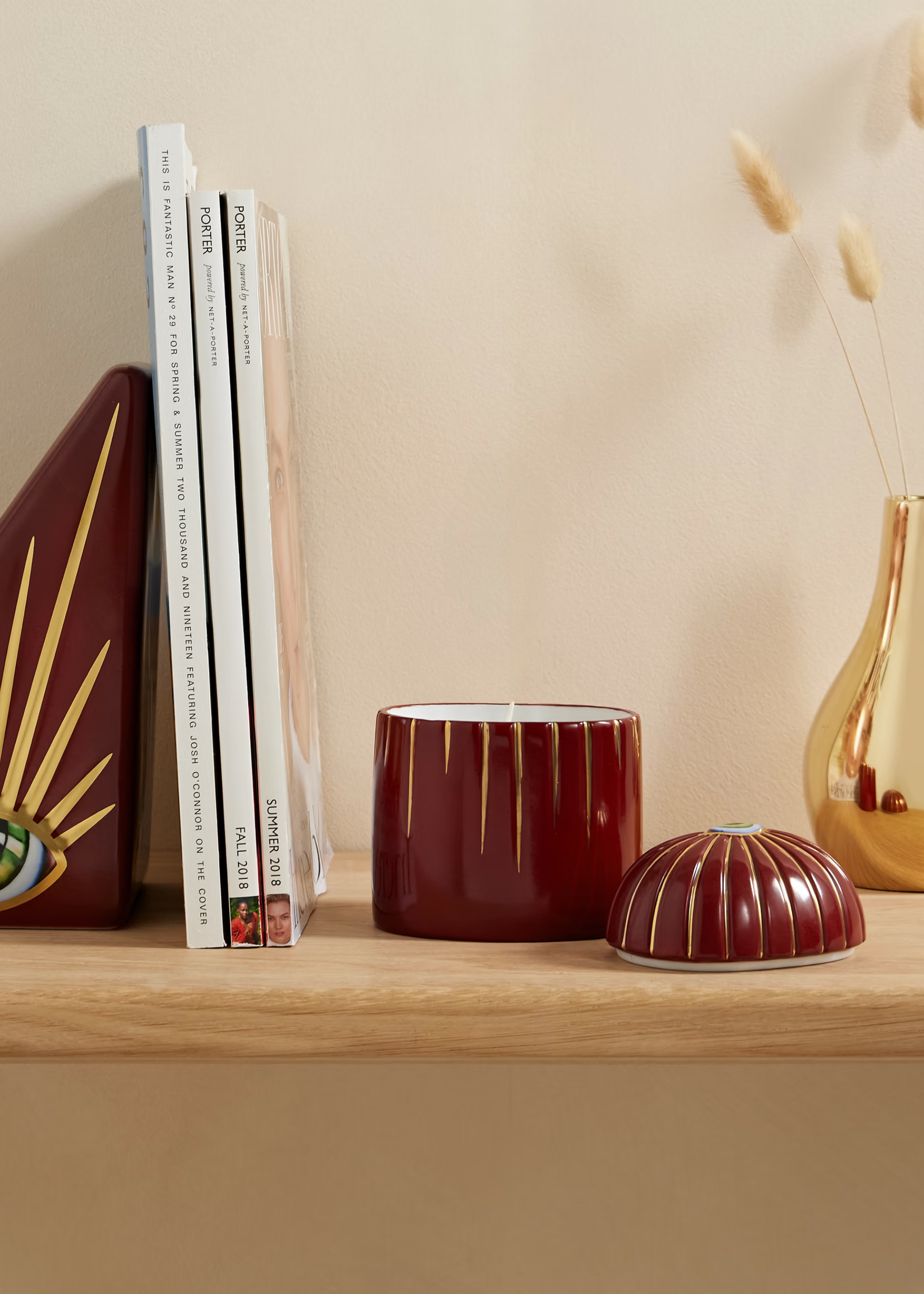 A floating light wood shelf with a stack of magazines, a red candle and lid beside a gold vase with dried stems