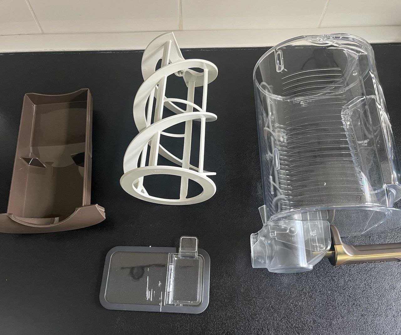 The individual compnants of the mixing unit of a Ninja Slushi Max, with the clear cannister, the brown condensation tray, the shite paddles, and the plastic lid, on a black kitchen counter.