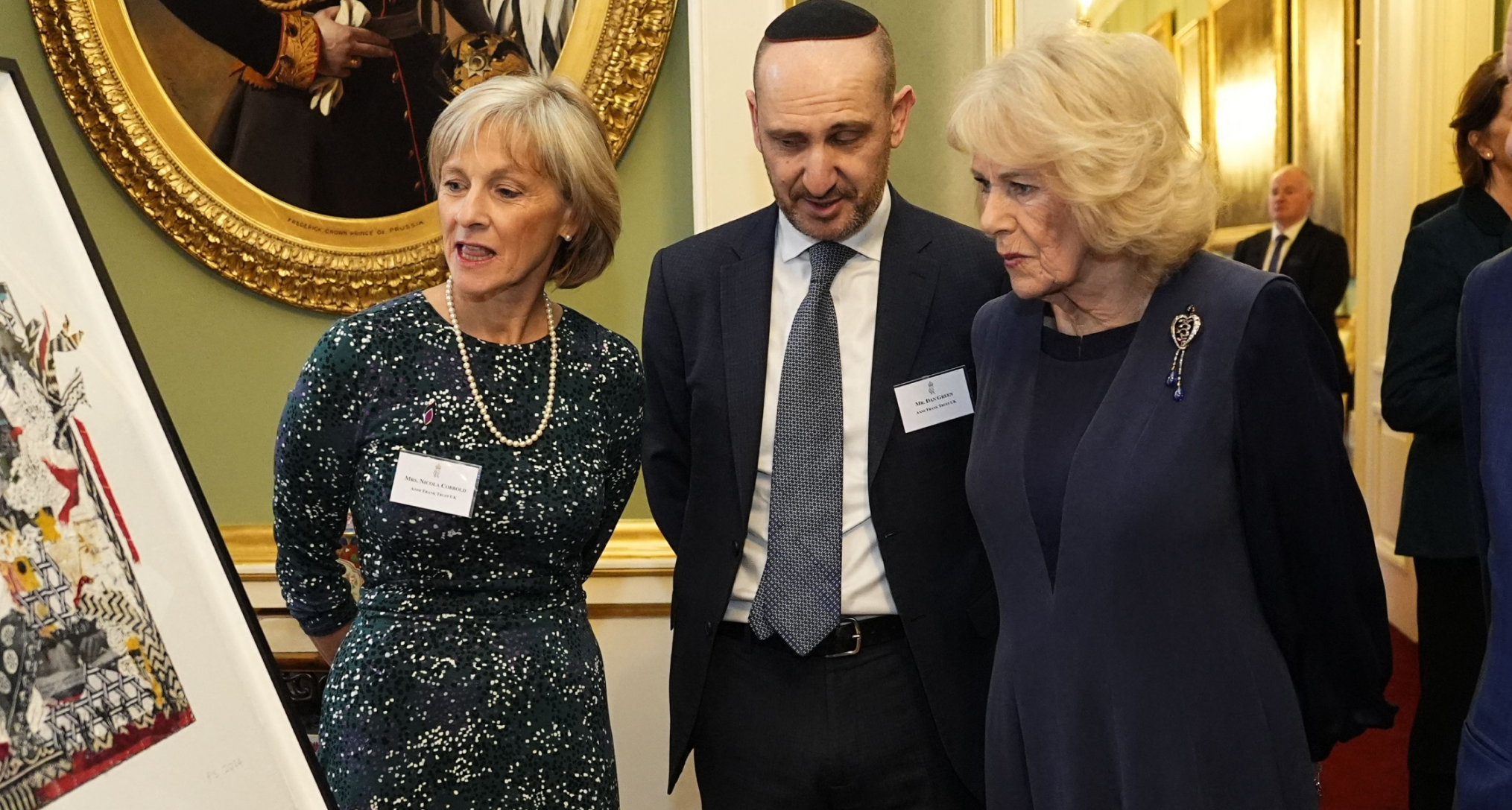 Queen Camilla wearing a blue jacket and a diamond heart brooch standing with a man and woman looking at a painting